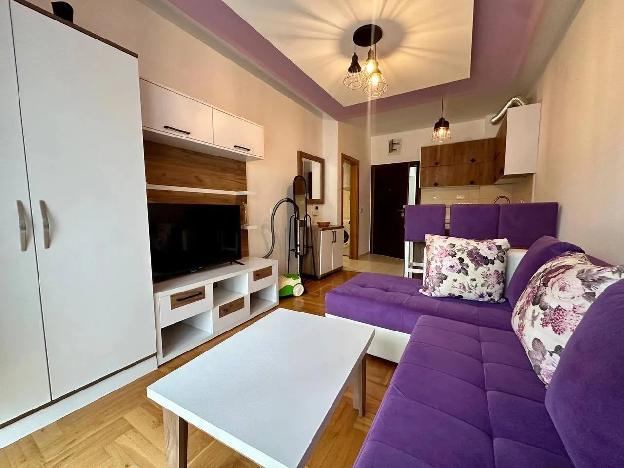 Living room with a sofa set, TV unit, and a kitchen in the background.