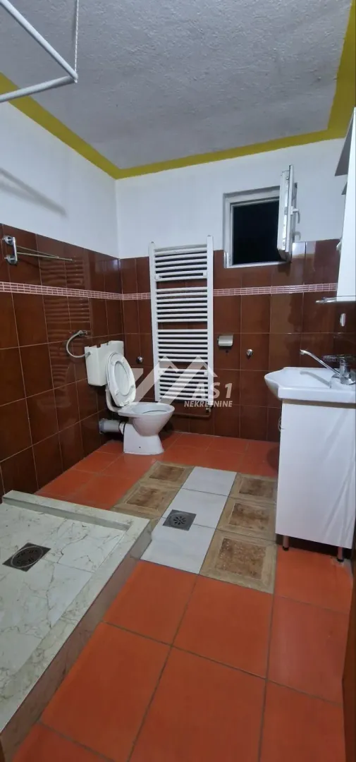 Kupatilo obloženo braon pločicama sa tuš kabinom, WC šoljom i umivaonikom.