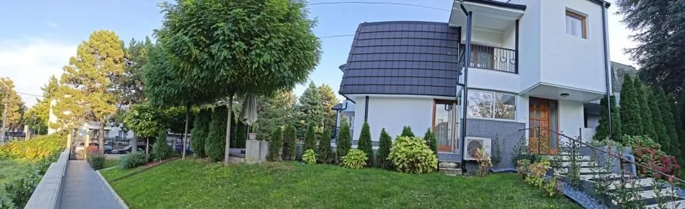 Pogled na dvorište i spoljašnjost kuće sa uređenim travnjakom i rastinjem.
