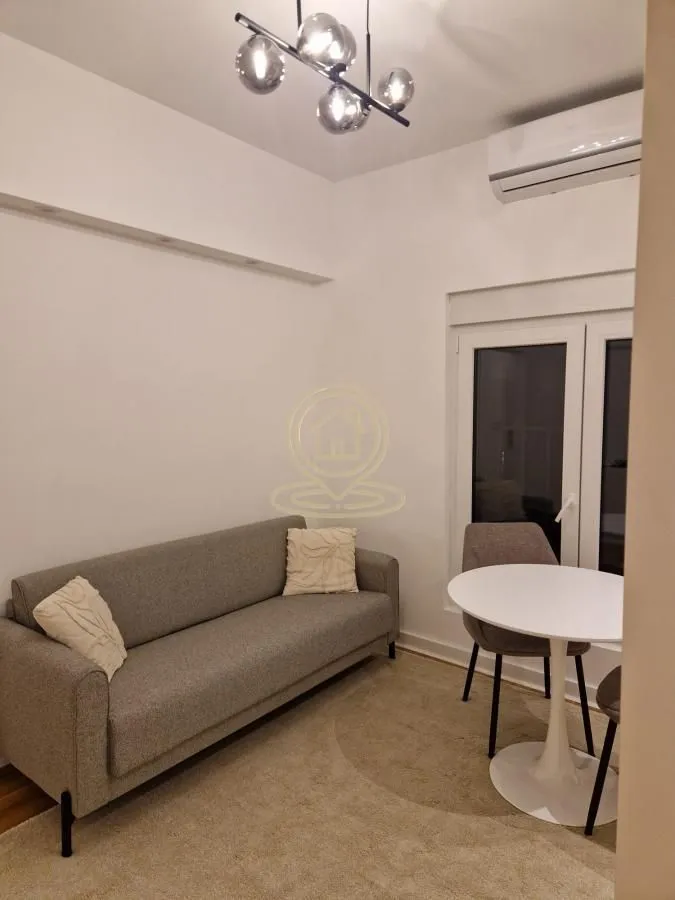 Manja dnevna soba sa sivim kaučem, trpezarijskim stolom i modernim lusterom.
