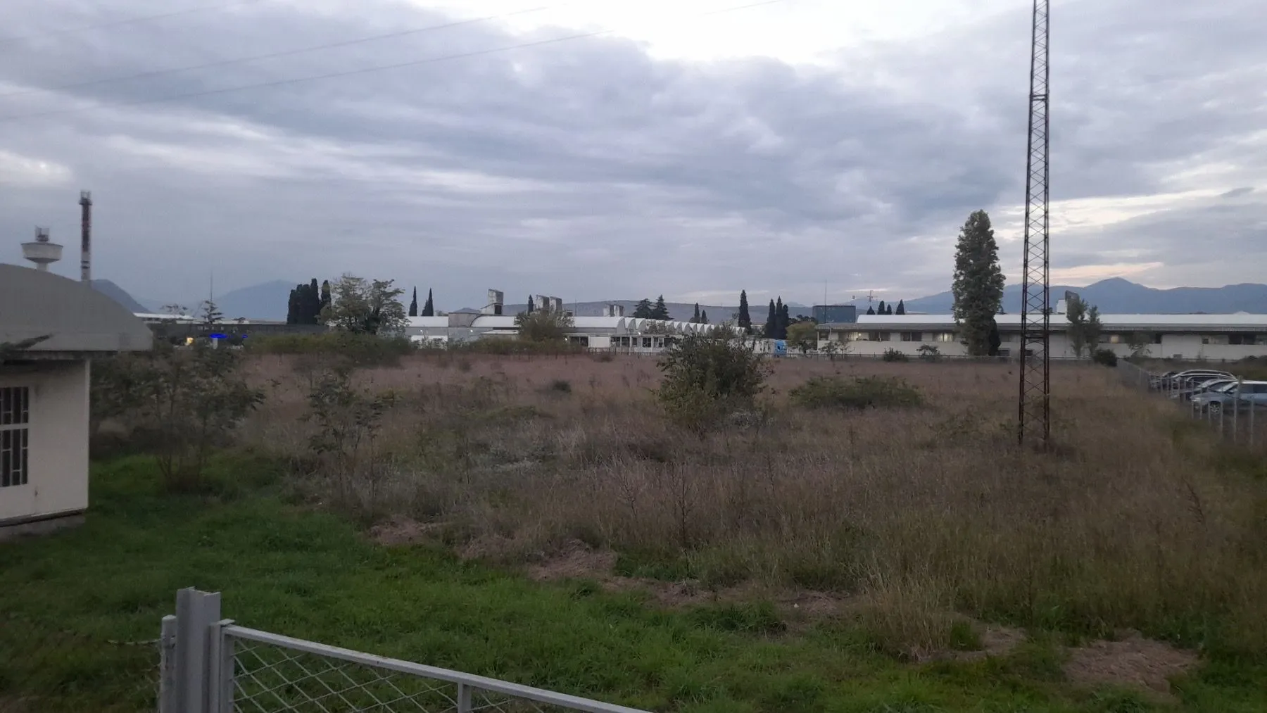 Pogled na prostrano zemljište sa niskim rastinjem i objektima u pozadini.