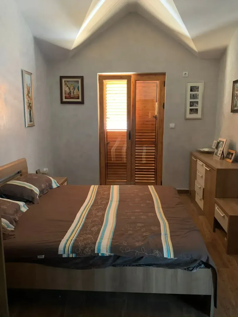 Spavaća soba sa bračnim krevetom, drvenim namještajem i balkonskim vratima.