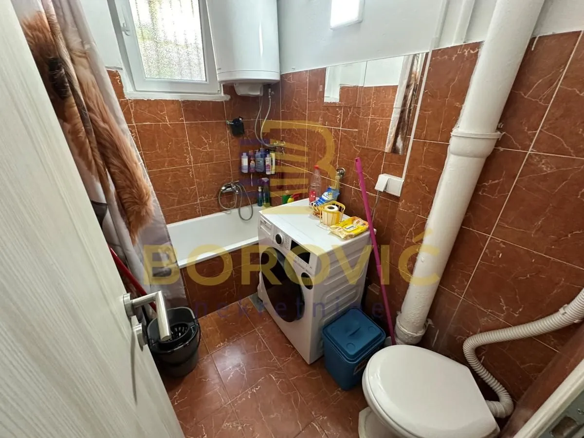 Manje kupatilo obloženo smeđim pločicama sa kadom, WC šoljom i veš mašinom.