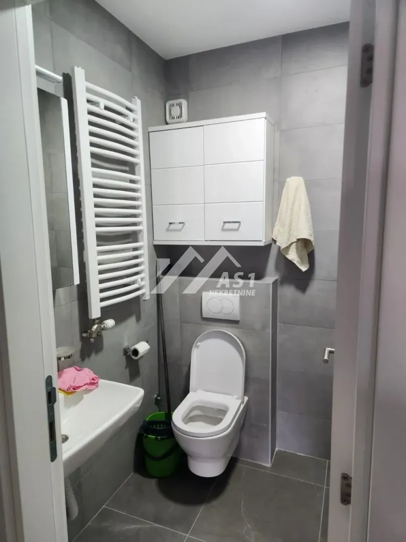 Manje kupatilo sa sivim pločicama, wc šoljom, lavaboom i belim ormarićem.