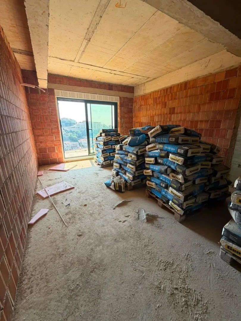 Prostorija u izgradnji sa gomilama građevinskog materijala i prozorom koji gleda na grad.