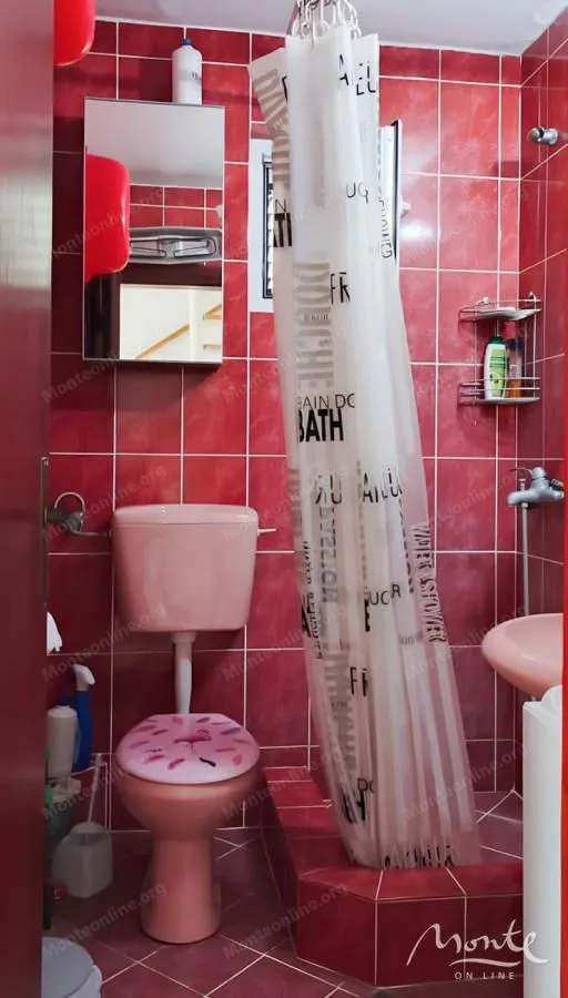 Kupatilo sa crvenim pločicama, wc šoljom, umivaonikom i tuš kabinom sa zavjesom.