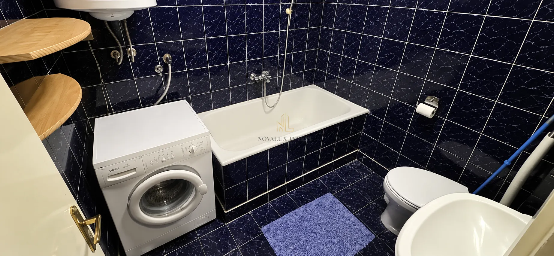 Kupatilo obloženo tamnim pločicama sa kadom, veš mašinom i sanitarijama.