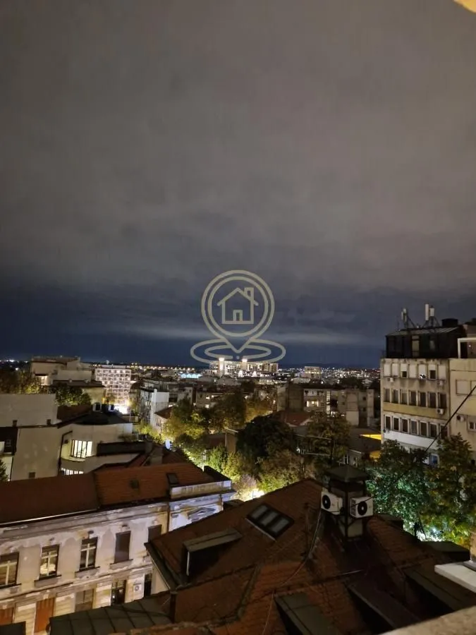 Noćni pogled sa prozora na gradske krovove i osvetljene ulice.