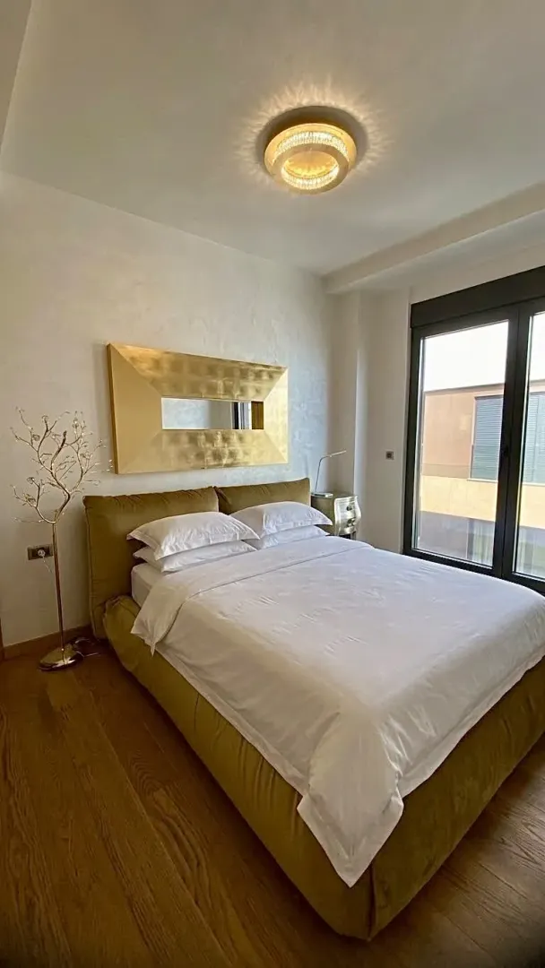 Bright bedroom featuring a double bed, a gold-framed mirror, and a large window.