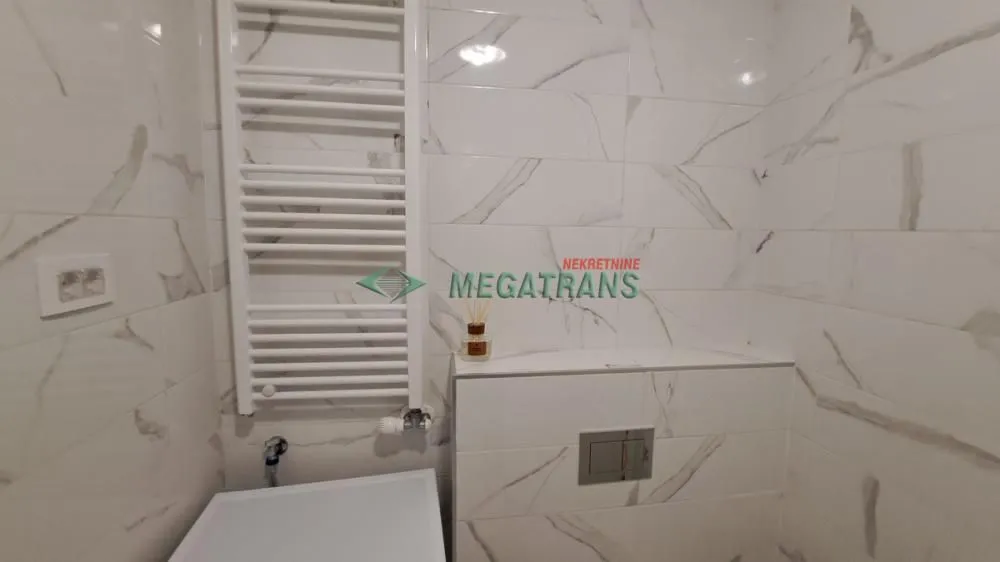 Kupatilo obloženo keramičkim pločicama sa mermernim dezenom i belim radijatorom.
