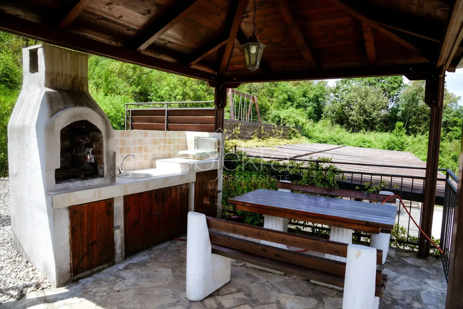 Natkriveni dio dvorišta sa roštiljem, sudoperom i drvenim stolom za sjedenje.