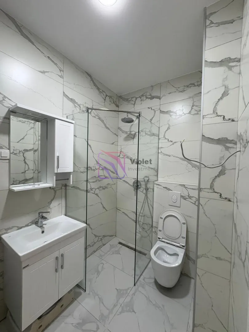 Moderne kupatilo sa tuš kabinom, umivaonikom i wc šoljom obloženo pločicama sa mermernim dezenom.