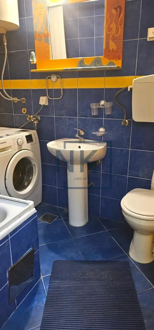 Kupatilo sa plavim pločicama, umivaonikom, WC šoljom i veš mašinom.