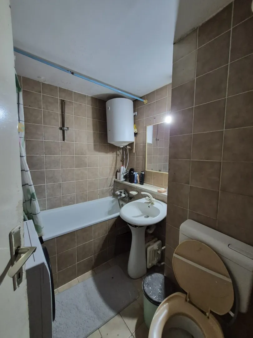 Manje kupatilo sa kadom, umivaonikom i wc šoljom obloženo braon pločicama.