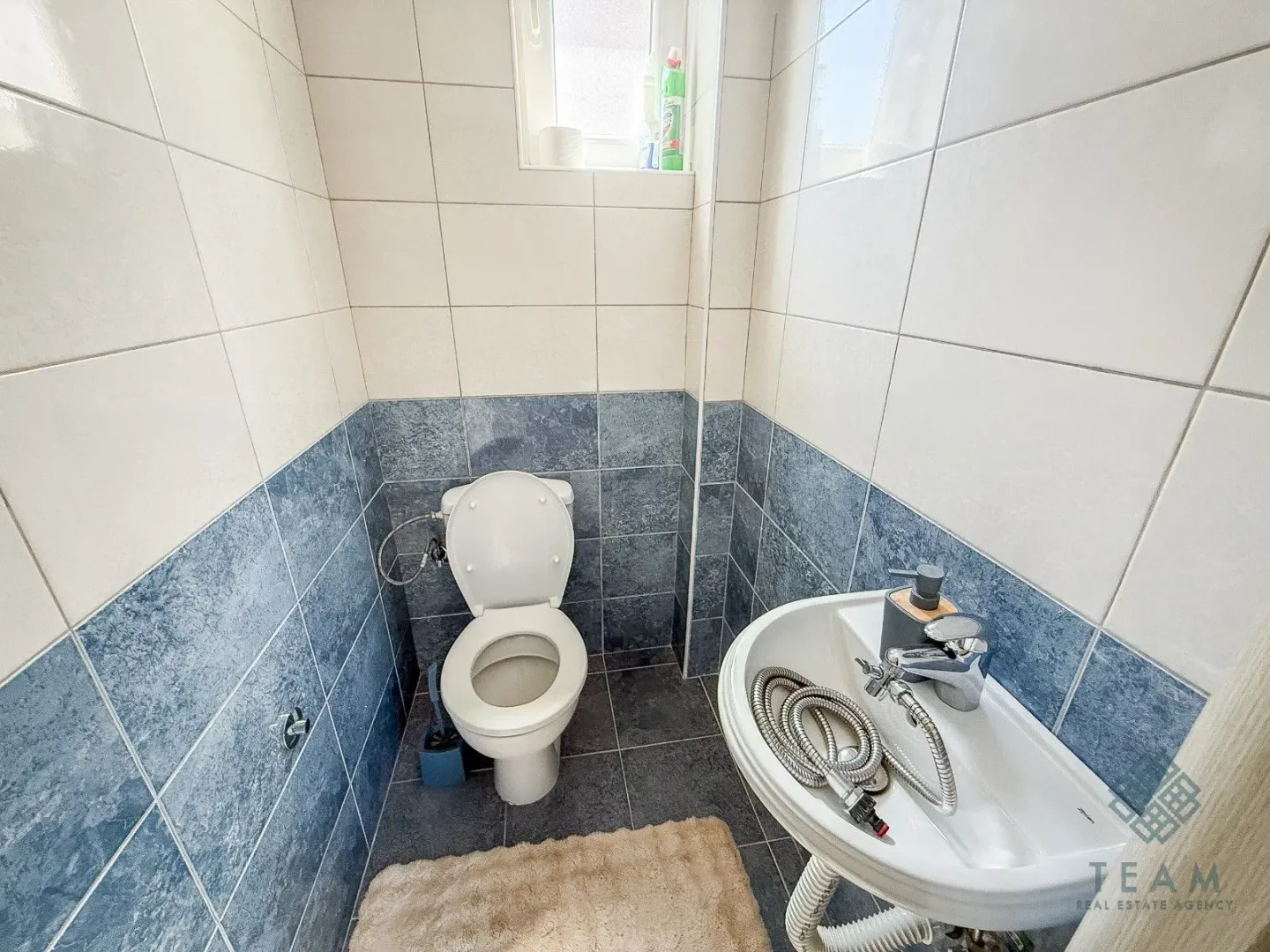 Manje kupatilo sa bijelim i plavim pločicama, wc šoljom i umivaonikom.