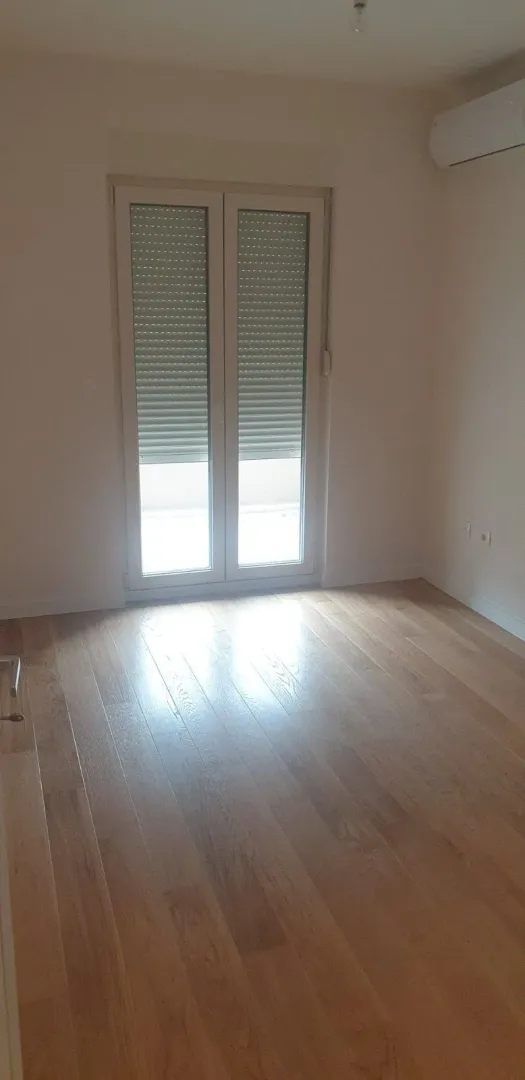 An empty room with light parquet flooring and balcony doors.