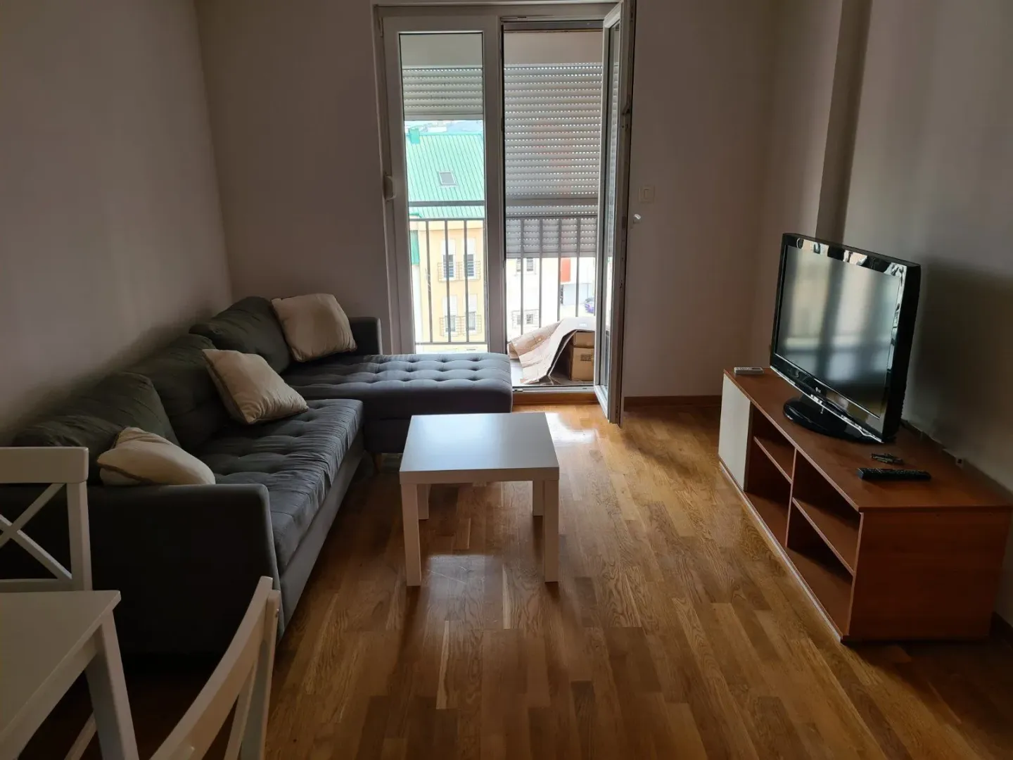 Svijetla dnevna soba sa sivom ugaonom garniturom, televizorom i izlazom na balkon.