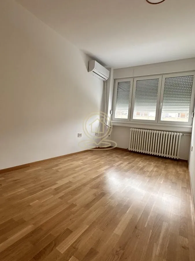 Prazna soba sa drvenim podom, klima uređajem i prozorom sa roletnama.