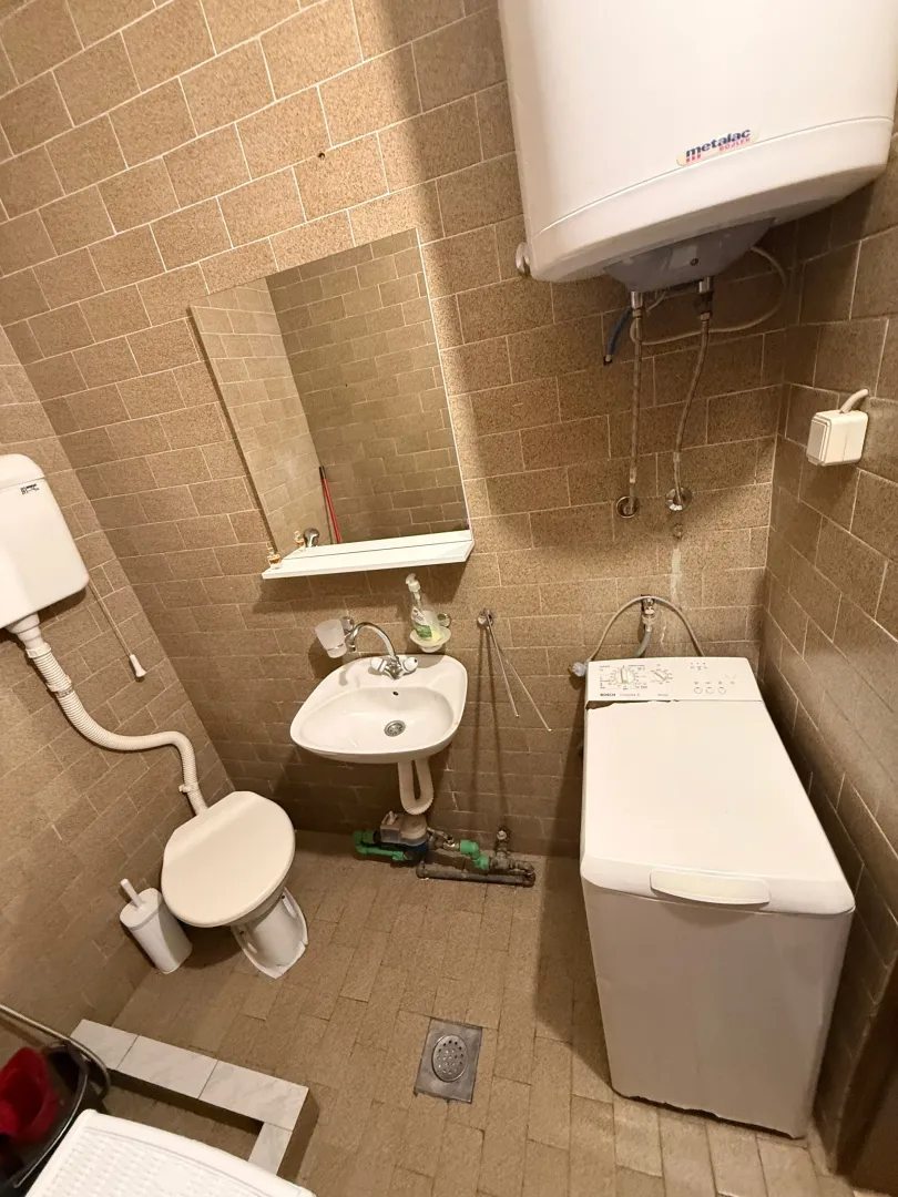 Manje kupatilo sa keramičkim pločicama, umivaonikom, WC šoljom i veš mašinom.