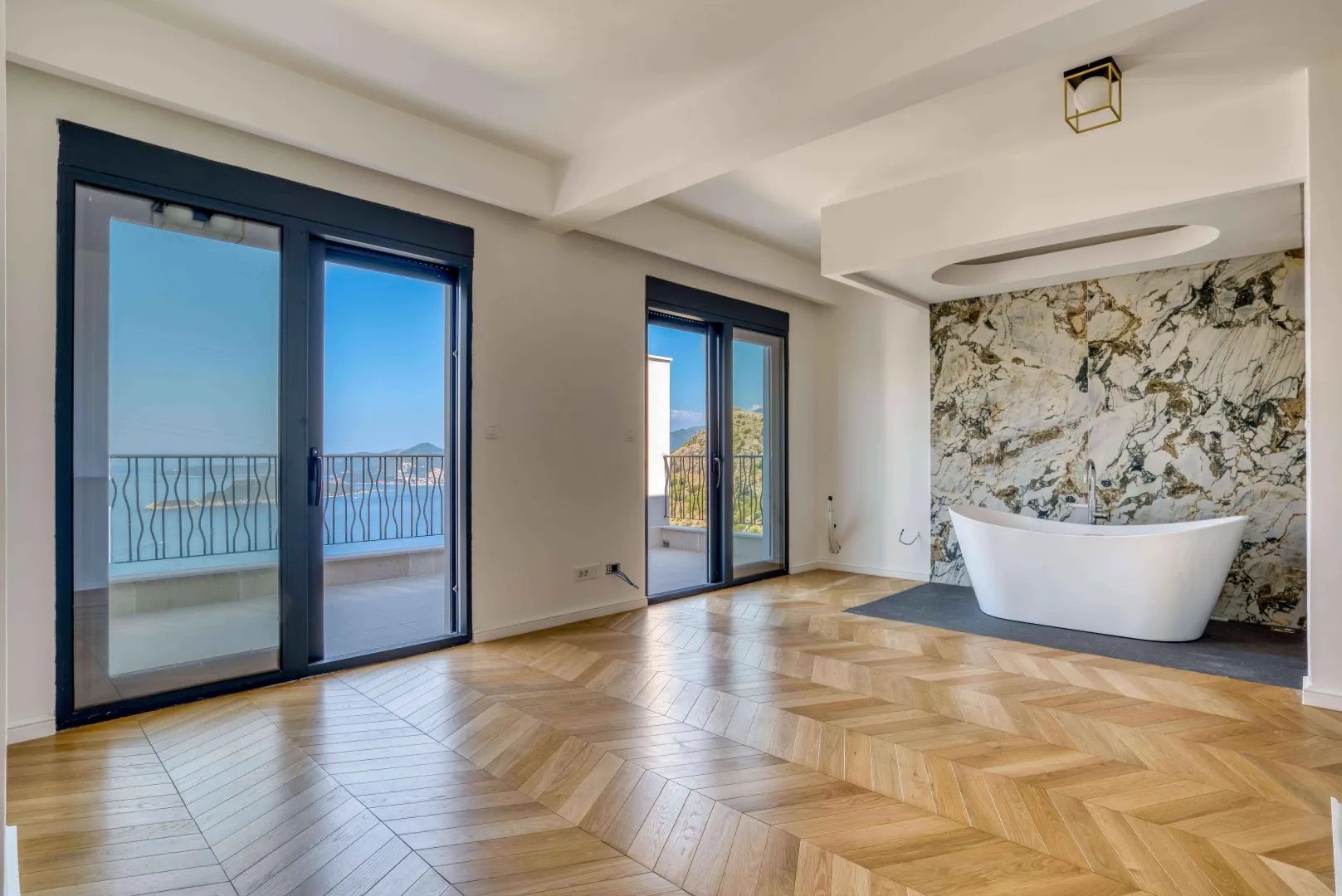 Prostrano kupatilo sa samostojećom kadom, drvenim podom i velikim staklenim vratima koja vode na balkon sa pogledom na more.