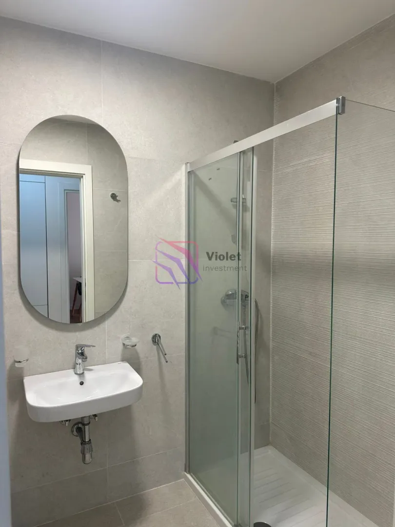A modern bathroom equipped with a sink, an oval mirror, and a walk-in shower.