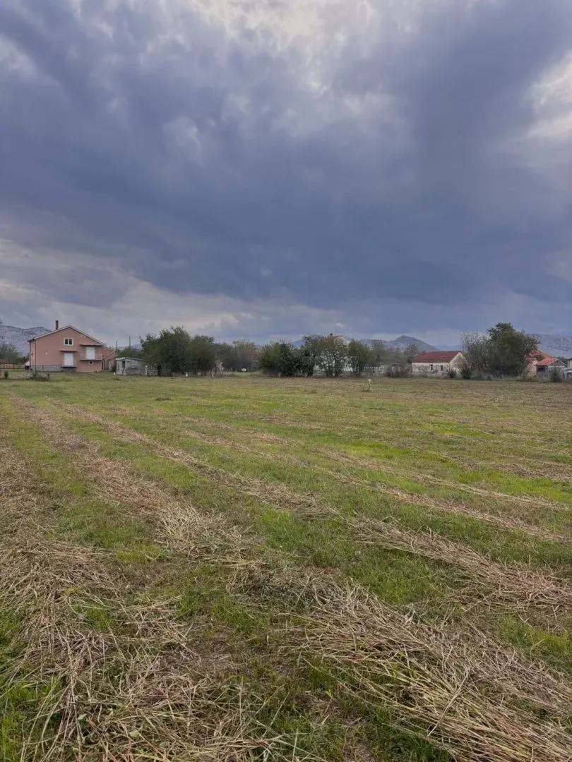 Pogled na prostrani plac sa travnatom površinom i okolnim kućama.