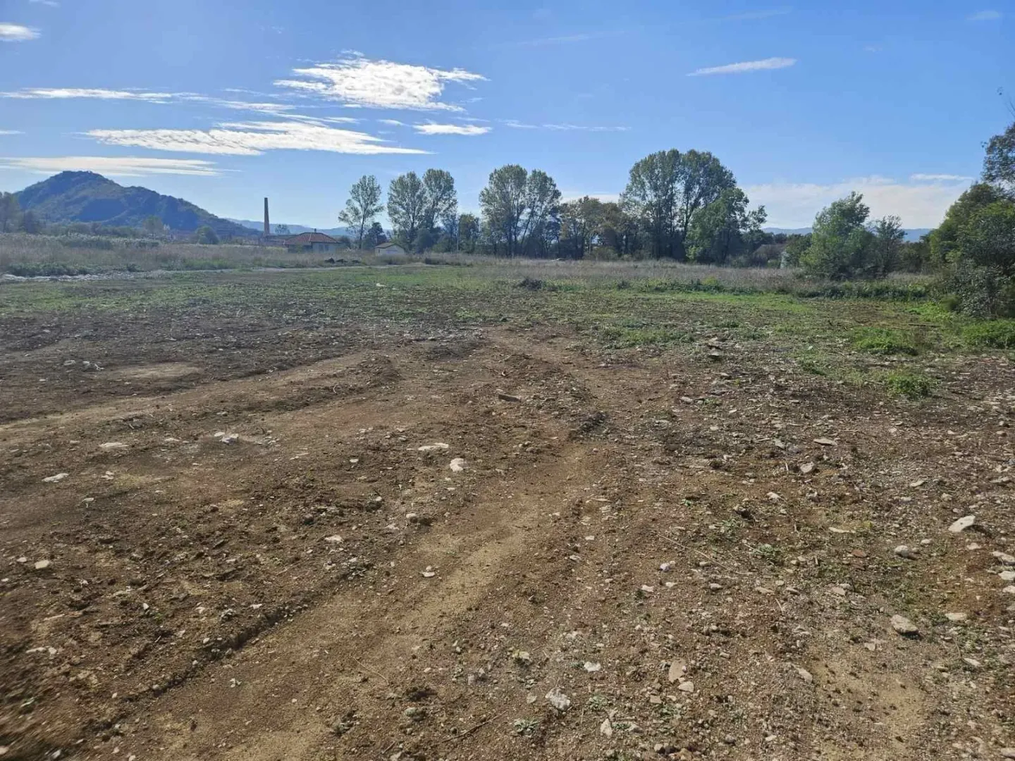 Prostran i ravan građevinski plac okružen drvećem pod vedrim nebom.