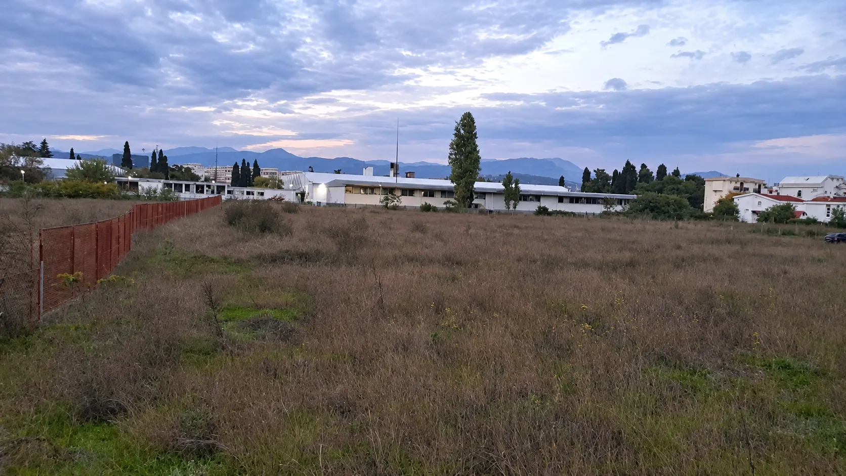 Pogled na prostrano zemljište sa ogradom i okolnim zgradama u daljini.