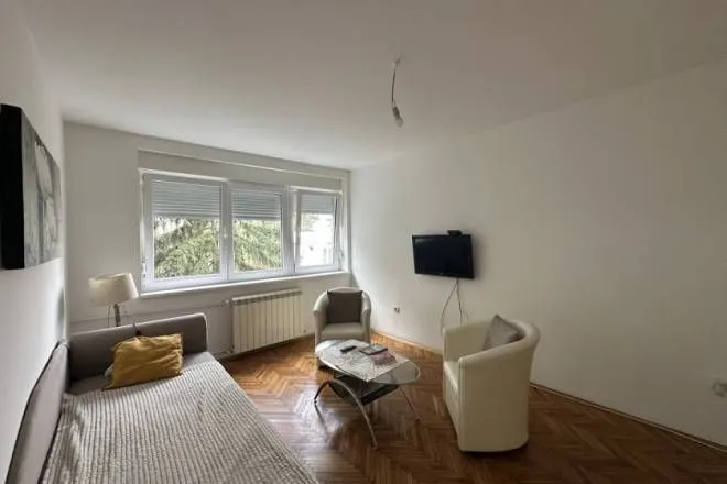 Dnevna soba sa krevetom, foteljama, stočićem i televizorom na zidu.