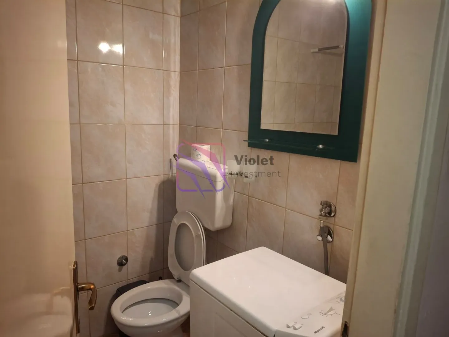 Manje kupatilo sa WC šoljom, mašinom za veš i ogledalom na zidu.