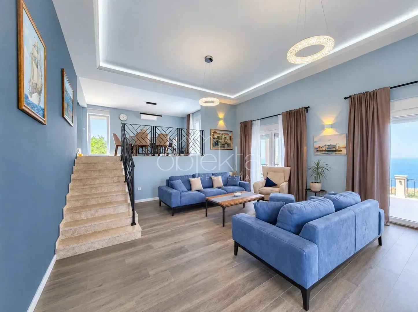Spacious living room with blue sofas, an elevated dining area and a staircase.