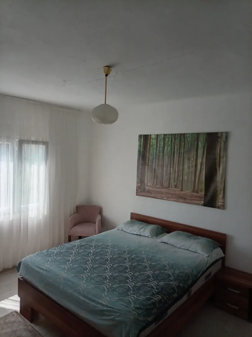 Spavaća soba sa bračnim krevetom, foteljom i prozorom sa zavjesama.