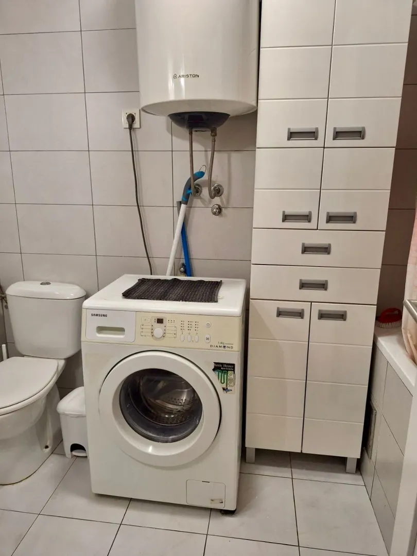 Kupatilo opremljeno WC šoljom, veš mašinom, bojlerom i bijelim ormarićem za odlaganje.