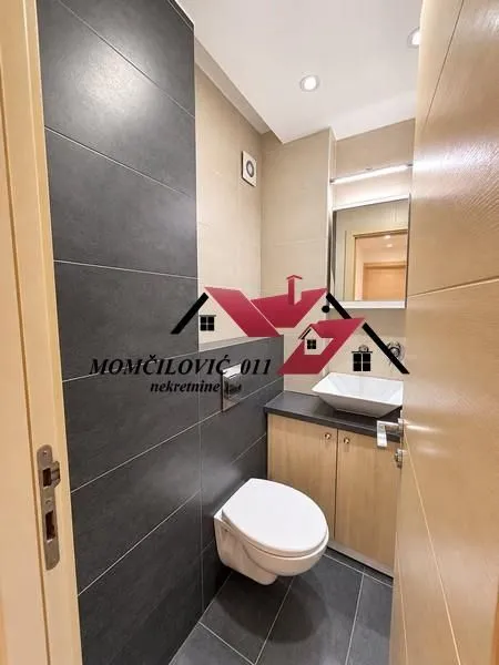 Manje kupatilo sa WC šoljom, umivaonikom i pločicama u tamnim tonovima.