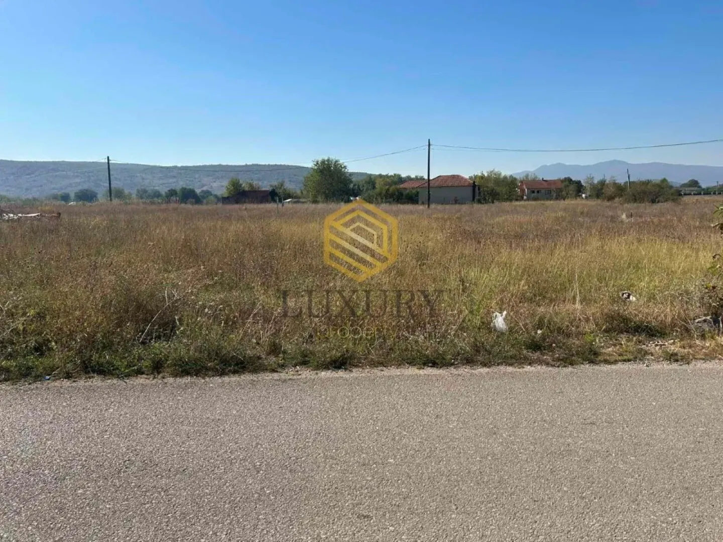 Pogled na ravno zemljište sa travom pored puta i kućama u pozadini.