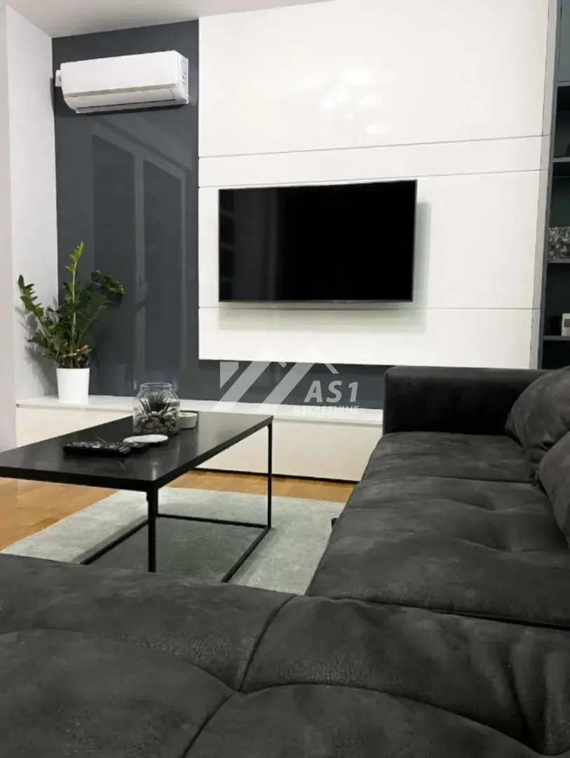 Dnevna soba sa ugaonom garniturom, stočićem i televizorom na zidu.