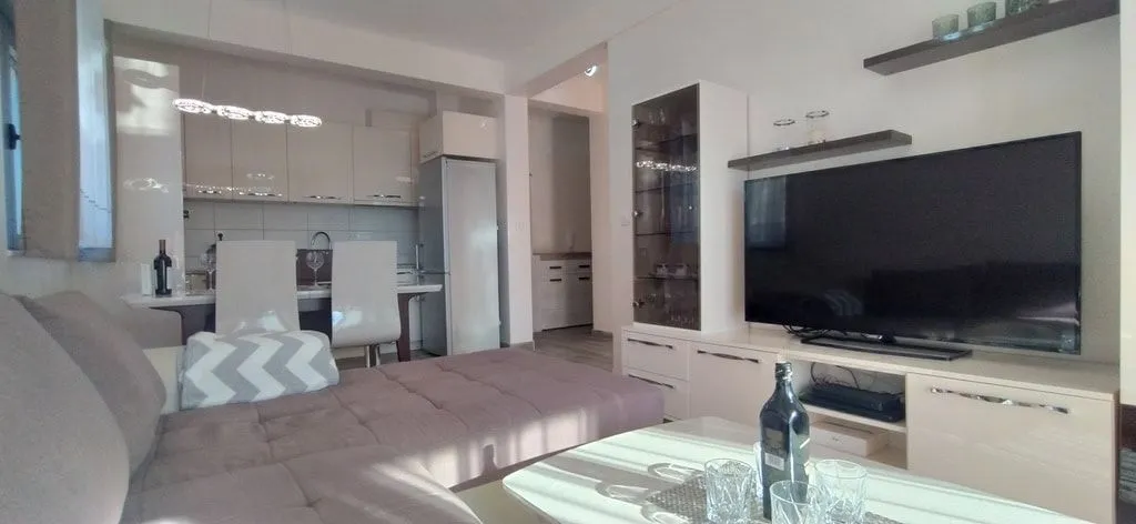 Svijetla dnevna soba sa ugaonom garniturom, televizorom i kuhinjom u pozadini.