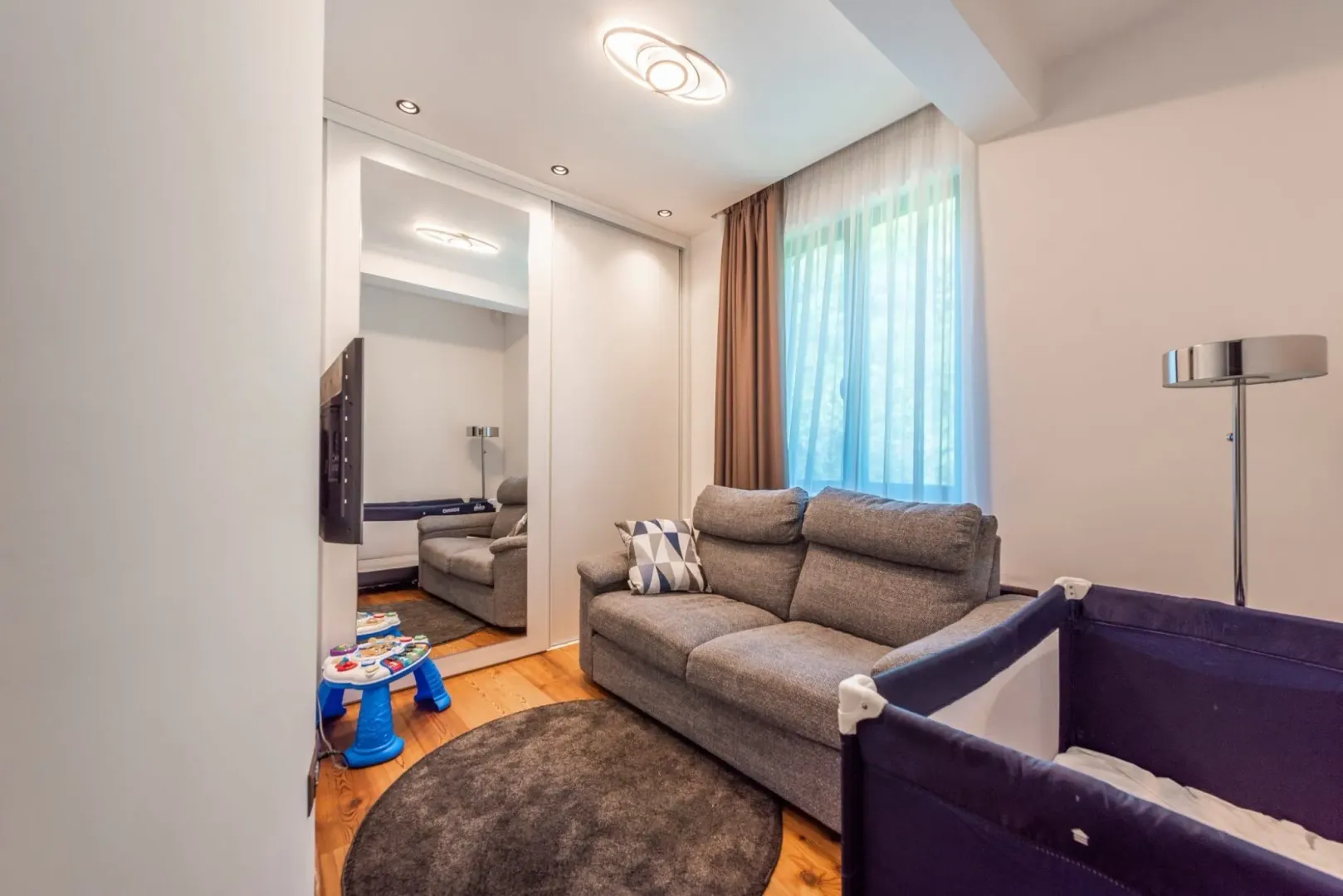 Bright room featuring a grey sofa, a baby crib, and a built-in wardrobe.