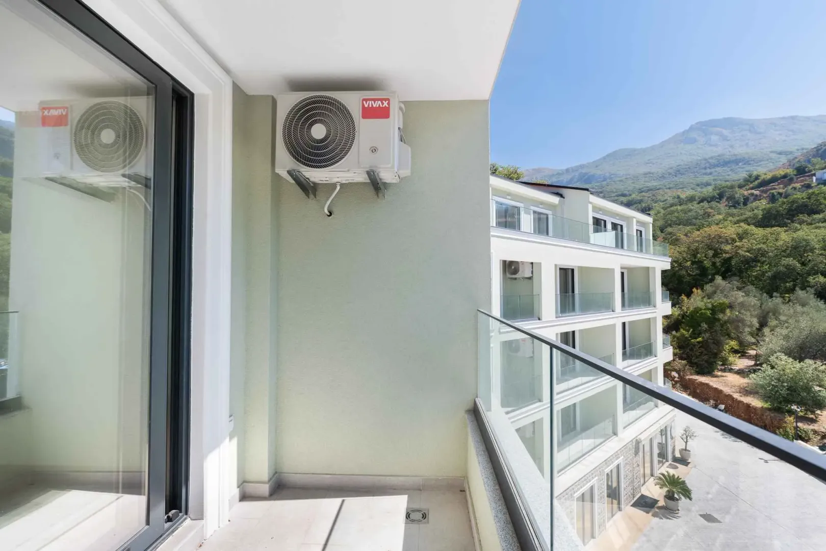 Manji svijetli balkon sa klima uređajem i pogledom na okolnu zgradu i brdo.