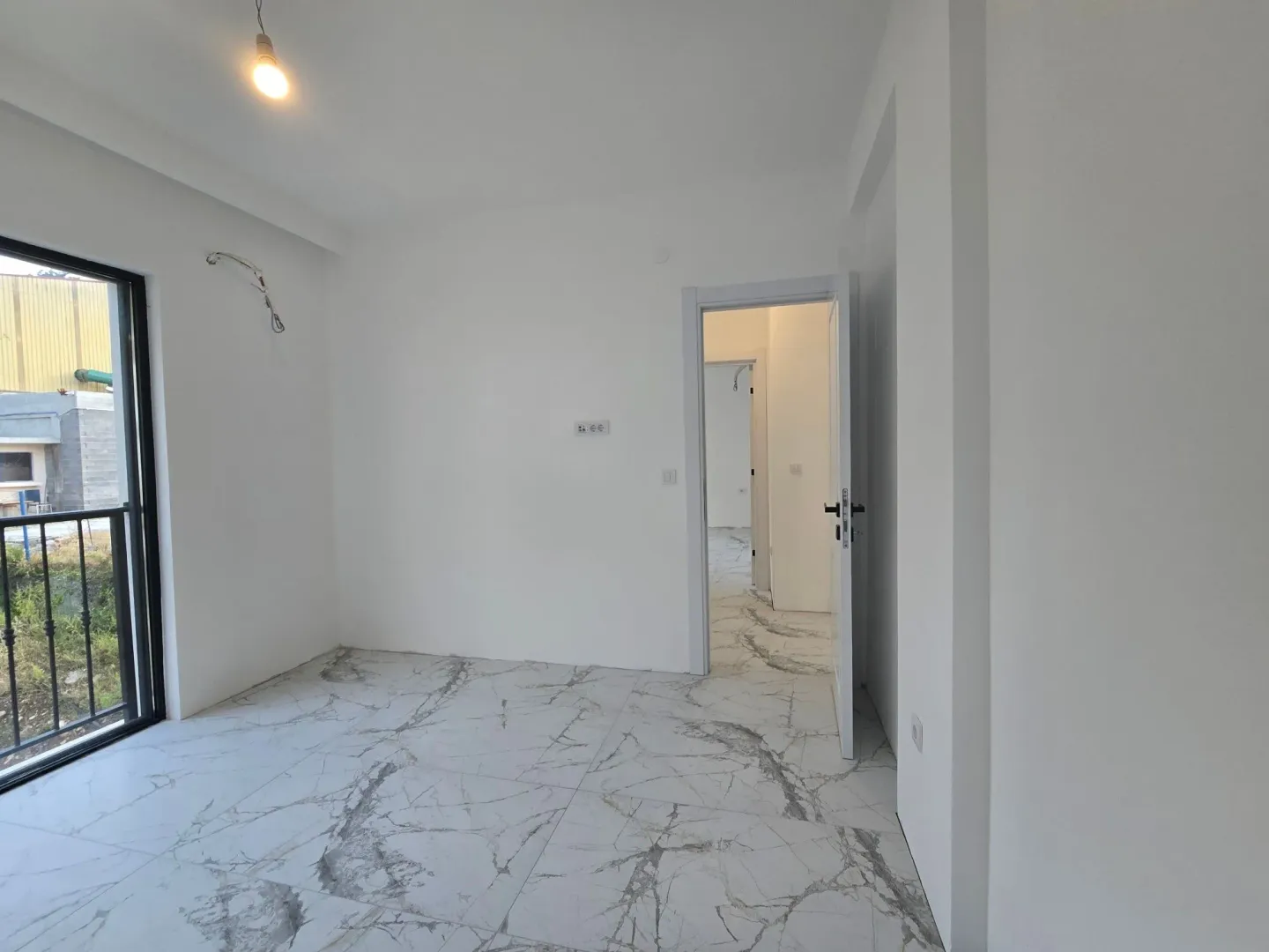 Prazna soba sa bijelim zidovima, mermernim pločicama na podu i izlazom na balkon.