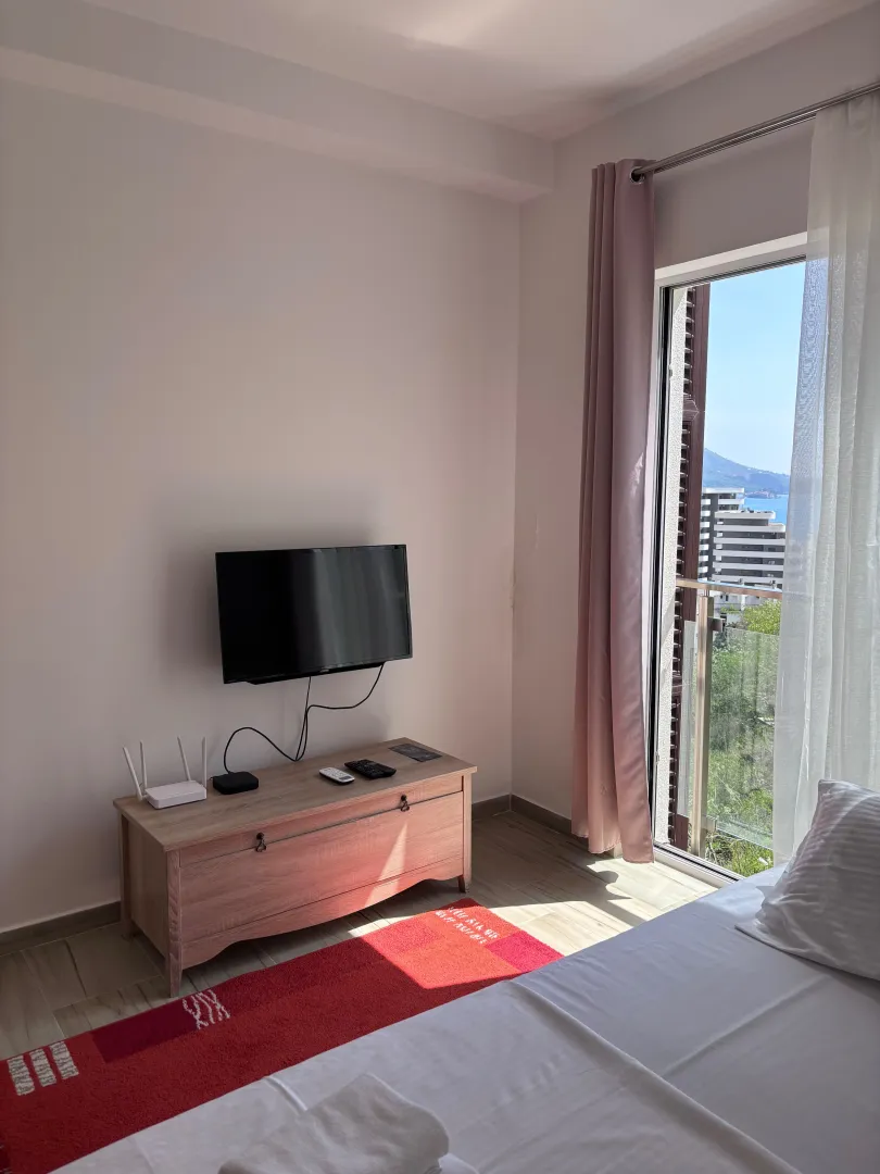 Svijetla spavaća soba sa krevetom, televizorom i izlazom na terasu sa pogledom na more.
