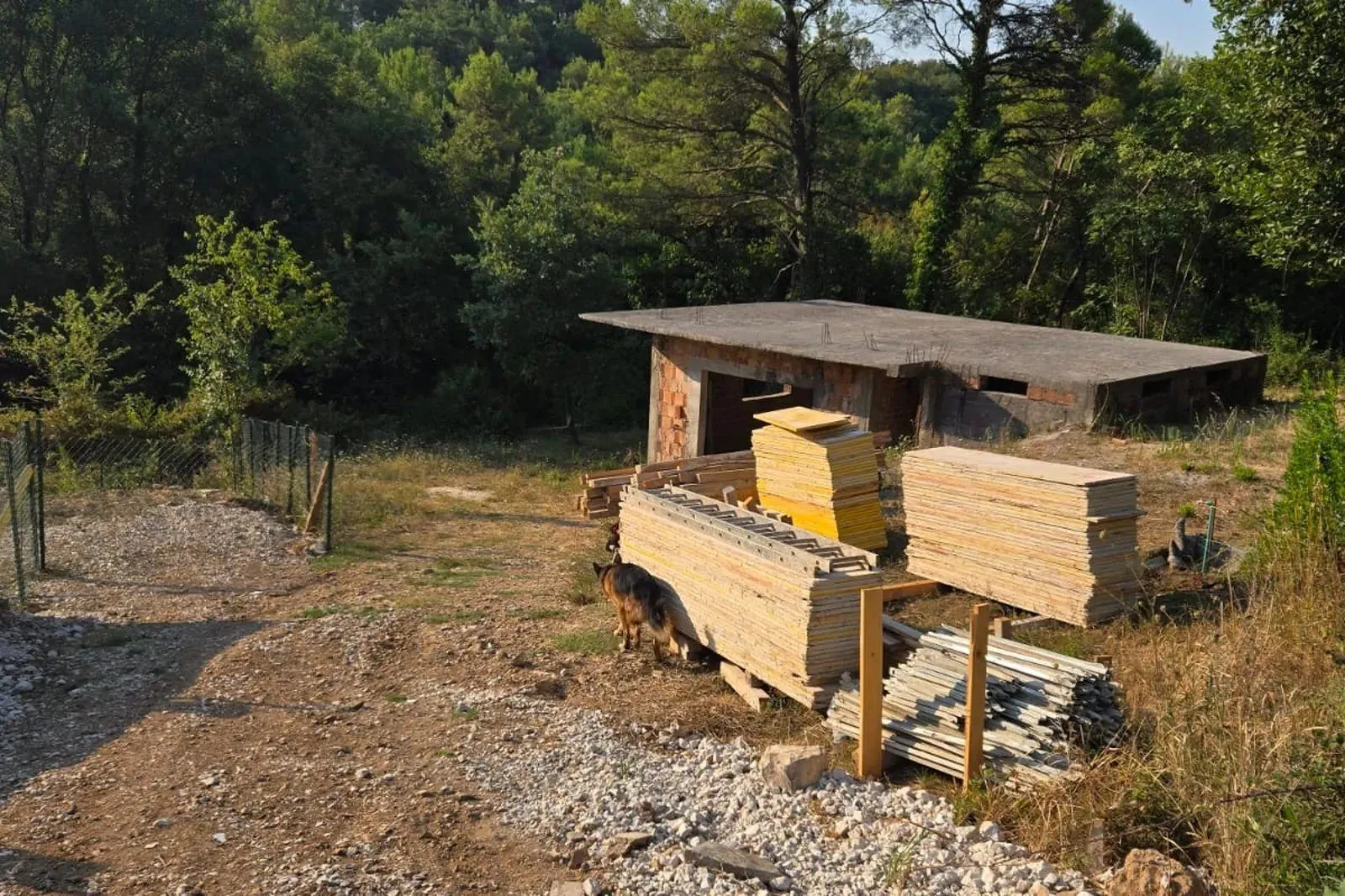 Nedovršeni objekat sa građevinskim materijalom u dvorištu okruženom šumom.