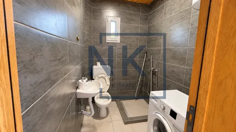 Kupatilo obloženo sivim pločicama sa tuš kabinom, WC šoljom, umivaonikom i veš mašinom.