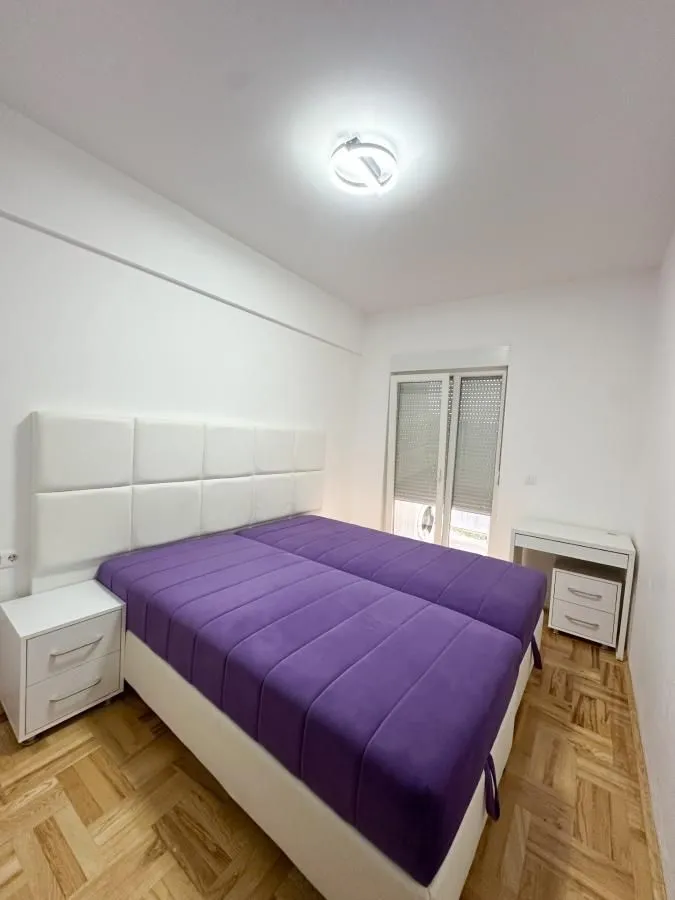 Spavaća soba sa velikim ljubičastim krevetom i belim noćnim stočićima.