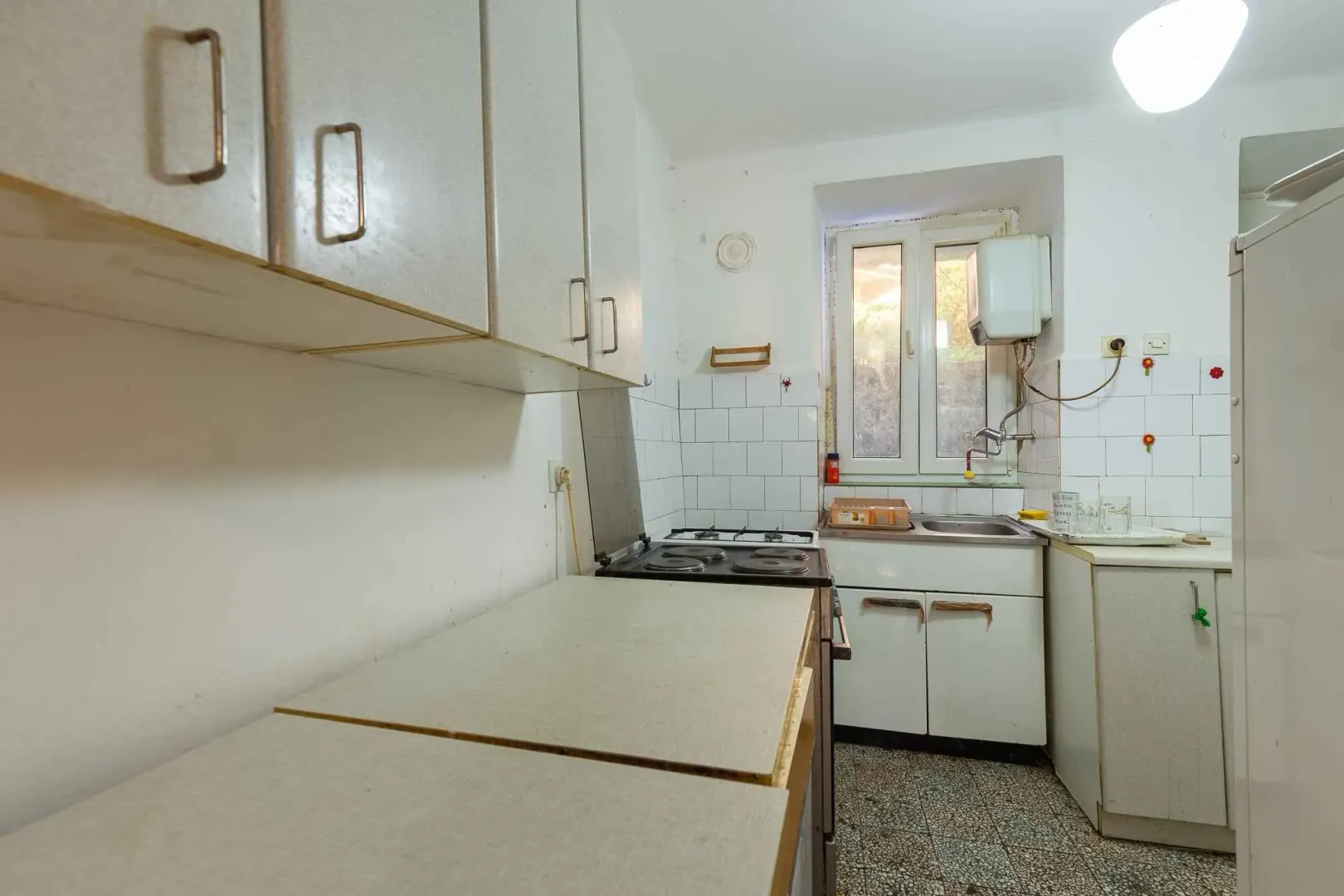 Starija kuhinja sa bijelim elementima, šporetom i manjim sudoperom pored prozora.