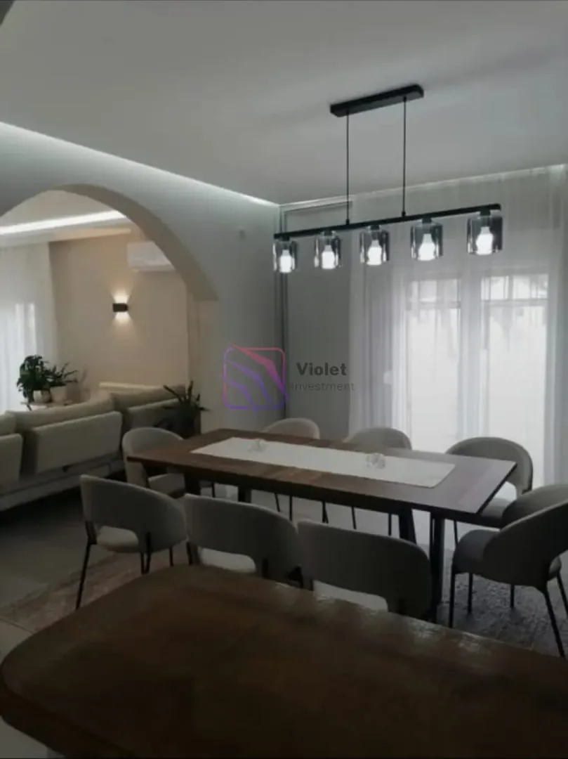 Dining area with a large table and modern lighting, with a view of the living room.