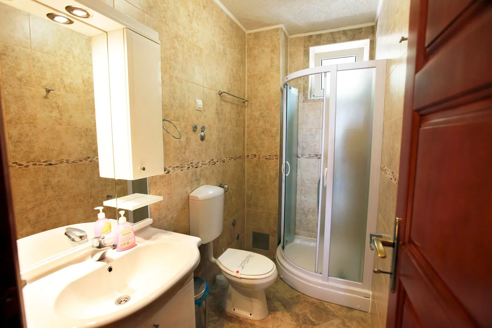 Smaller bathroom with a shower cabin, sink, and toilet in beige tiles.
