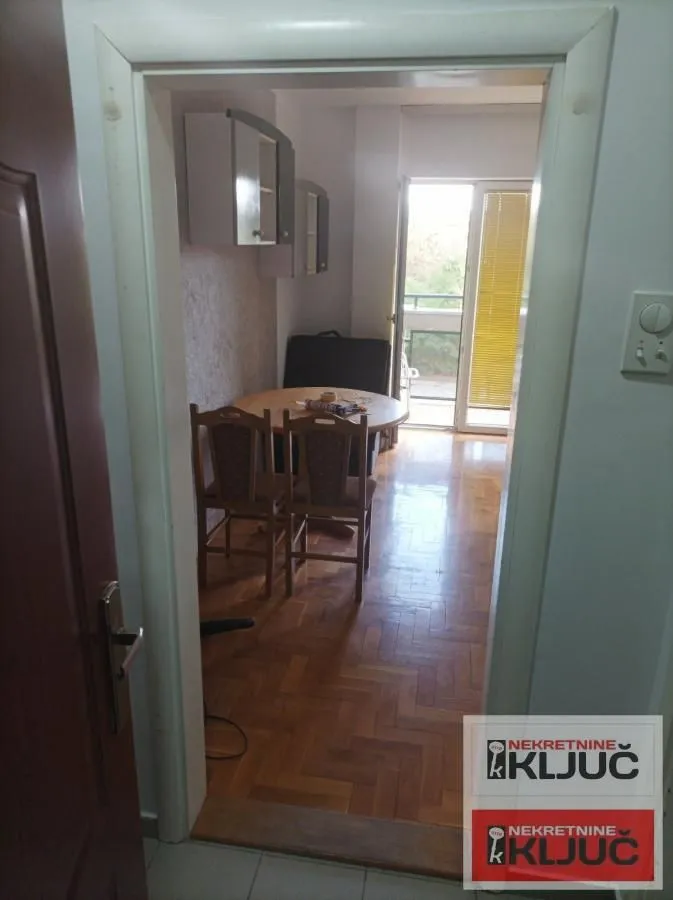 Pogled iz hodnika na trpezariju sa stolom, stolicama i kuhinjskim elementima.
