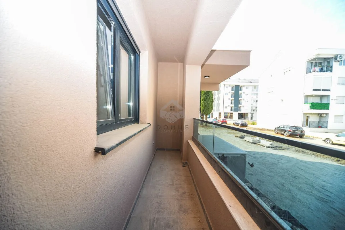 Natkriveni balkon sa staklenom ogradom i pogledom na okolne zgrade i parking.